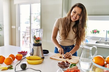 Young beautiful hispanic woman preparing vegetable smoothie with blender cutting date at the kitchen