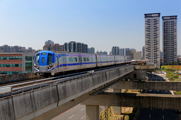 A metro train travels on the elevated rails of Taoyuan Airport MRT System with residential tower blocks in background under blue sunny sky in Chunli, Taoyuan, Taiwan
