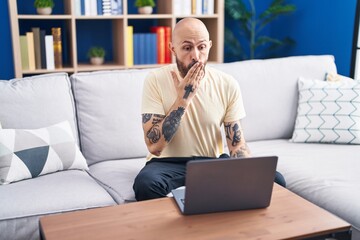 Hispanic man with tattoos using laptop at home covering mouth with hand, shocked and afraid for...