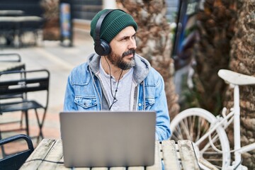 Young bald man using laptop and headphones sitting on table at coffee shop terrace