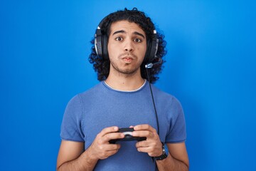 Hispanic man with curly hair playing video game holding controller puffing cheeks with funny face. mouth inflated with air, catching air. - obrazy, fototapety, plakaty