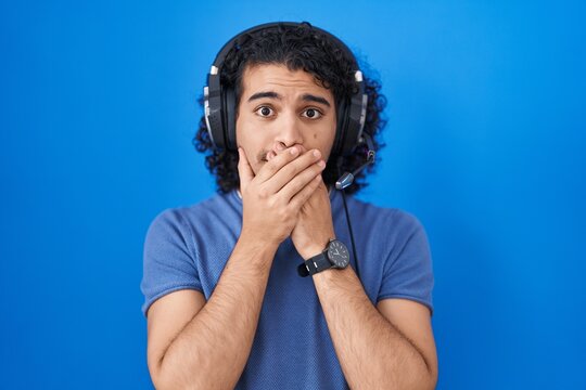 Hispanic man with curly hair listening to music using headphones shocked covering mouth with hands for mistake. secret concept.