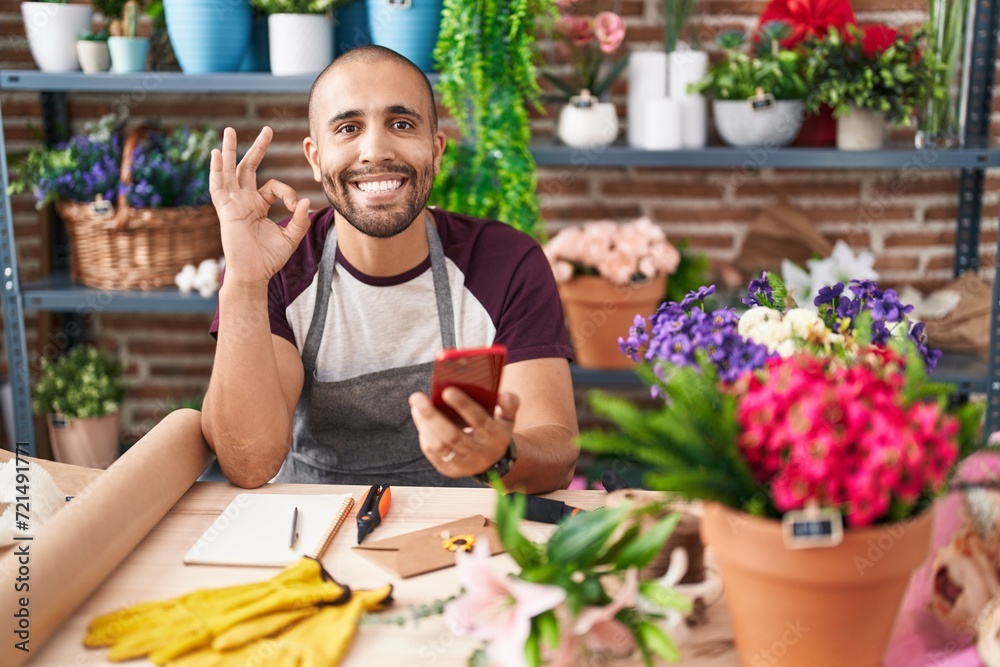 Poster Hispanic man with beard working at florist shop with smartphone doing ok sign with fingers, smiling friendly gesturing excellent symbol