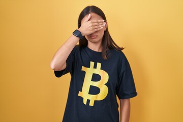 Young hispanic woman wearing bitcoin t shirt covering eyes with hand, looking serious and sad....