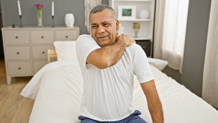 Obraz premium Mature hispanic man in bedroom setting displaying discomfort or soreness, holding his neck.
