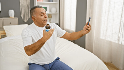 Mature hispanic man checking blood sugar level with glucometer while sitting on bed in a bedroom.