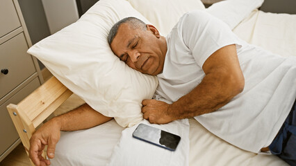 Mature hispanic man sleeping peacefully in a bedroom, with a smartphone by his side.