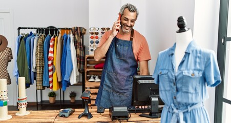 Middle age grey-haired man shop assistant using computer talking on smartphone at clothing store