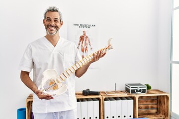 Middle age grey-haired man physiotherapist smiling confident holding anatomical model of spinal...
