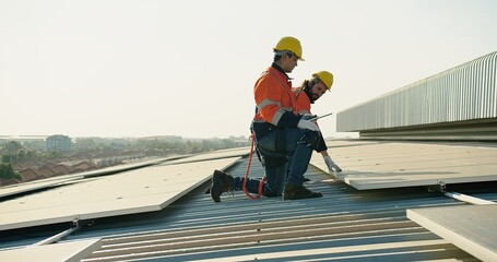 Two engineer workers in safety gear install solar panels on a rooftop, with a suburban landscape...