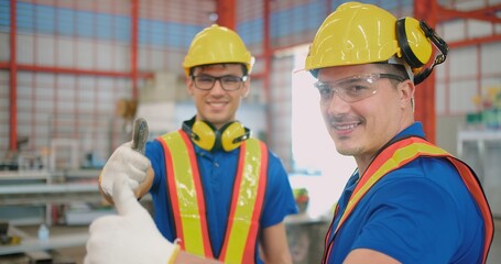 Two smiling Engineers construction industrial workers wearing hardhats safety gear giving thumbs up inside an industrial manufacturing factory
