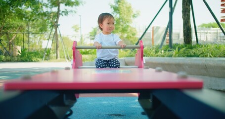 Front view of a focused curious toddler sits on a seesaw, hands on the handlebar with playground...