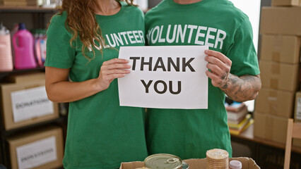 A man and woman in green 'volunteer' t-shirts holding a 'thank you' sign indoors with donation...