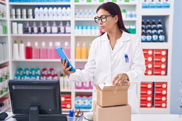 Young beautiful hispanic woman pharmacist using touchpad holding pills bottle at pharmacy