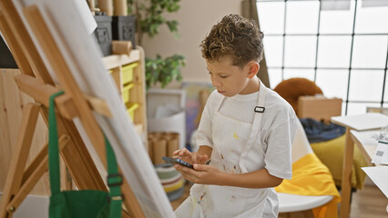 Adorable blond boy artist earnestly looking at smartphone, learning to draw in cozy art studio class