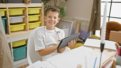 Adorable blond boy artist confidently drawing on touchpad in art studio classroom, smiling as he...