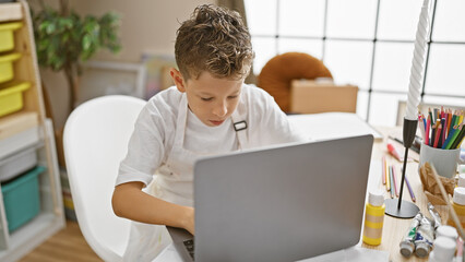 Adorable blond artist boy seriously concentrating on laptop at art studio, sitting in a cute apron,...