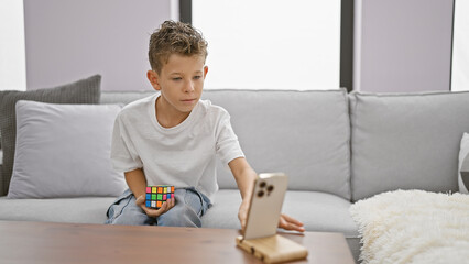 Adorable blond boy confidently solving rubik's cube puzzle, playing with smartphone turning on,...