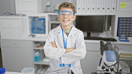 Confident and smiling blond boy scientist in lab, cute kid with arms crossed in adorable gesture, sitting at work table, radiating happy confidence - obrazy, fototapety, plakaty