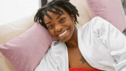 A cheerful young african american woman with dreadlocks lying on a bed indoors, wearing a white...