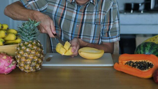 Young man cutting resh ripe mango fruit on a chopping board, slow motion. Lots of exotic tropical organic fruits are on the table. Healthy lifestyle and natural organic food concept