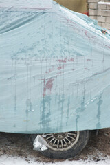 An old car stands on the street in the open air, covered with a protective tarpaulin, oilcloth, or cover to protect it from snow and rain in winter. Close-up photo.