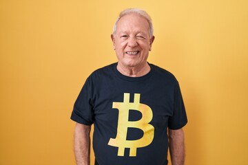 Senior man with grey hair wearing bitcoin t shirt winking looking at the camera with sexy...