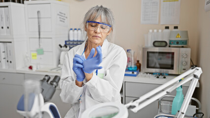 A mature woman scientist in a laboratory setting is putting on gloves for an experiment, surrounded by equipment and chemicals.