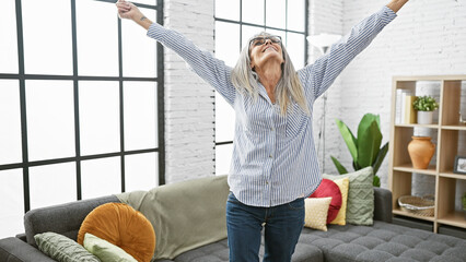 Mature woman stretching with joy in a cozy living room with sofa, plants, and a large window.