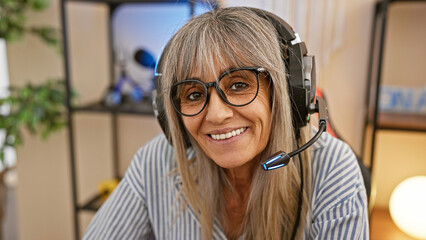 Smiling mature woman with grey hair wearing a headset in a cozy indoor office space at night.