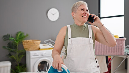 Middle age grey-haired woman talking on smartphone ironing clothes at laundry room