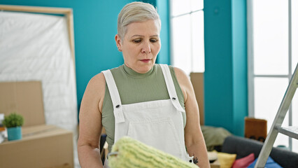 Middle age grey-haired woman holding paint roller with serious face at new home
