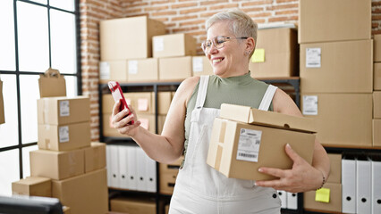 Middle age grey-haired woman ecommerce business worker using smartphone holding package at office