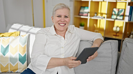 Middle age grey-haired woman using touchpad sitting on sofa at home