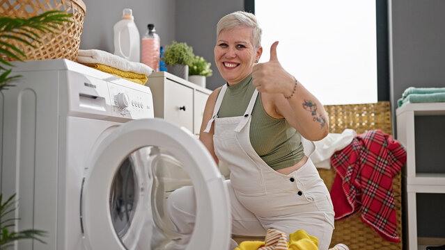 Middle age grey-haired woman smiling confident washing clothes doing thumb up gesture at laundry room