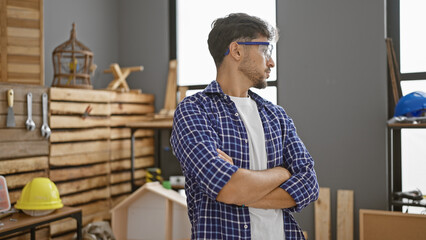 Serious-faced young arab carpenter with crossed arms standing formidably in his carpentry workshop, exemplifying the handsome adult male woodworker in the furniture industry