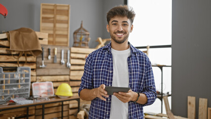 Smiling young arab man takes on carpentry business, embracing technology with touchpad in his...