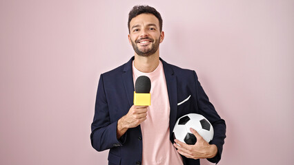 Young hispanic man soccer reporter holding microphone and ball smiling over isolated pink background