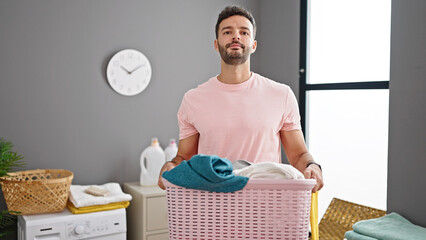 Young hispanic man holding basket with clothes at laundry room