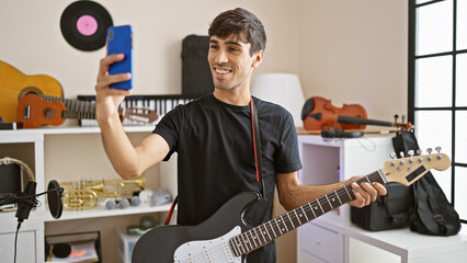 Smiling young hispanic man, a rockstar musician, electrifies the room making a selfie with his electric guitar in a music studio