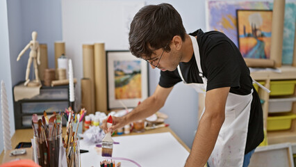 Young, handsome hispanic man seriously concentrating on drawing art in studio, brushes and palette at the ready