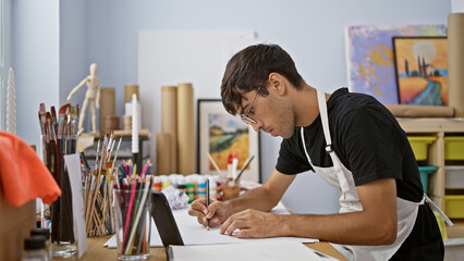 Attractive young hispanic man ardently draws in art studio, engrossed in his smartphone, surrounded by brushes and paint palette
