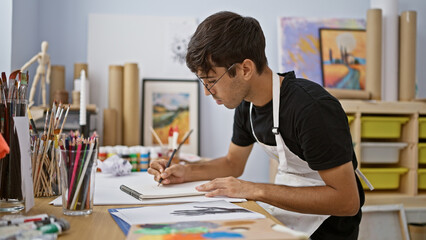 Talented young hispanic artist fully absorbed in sketching with pencil at a lively art studio,...