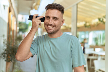 Young hispanic man smiling confident listening audio message by the smartphone at coffee shop terrace