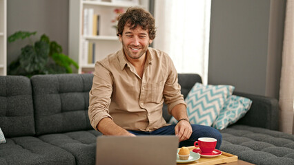 Young hispanic man having breakfast using laptop smiling at home - Powered by Adobe