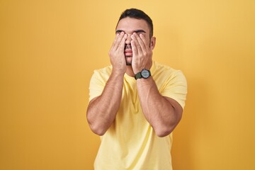 Young hispanic man standing over yellow background rubbing eyes for fatigue and headache, sleepy...