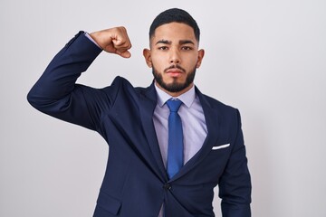 Young hispanic man wearing business suit and tie strong person showing arm muscle, confident and proud of power