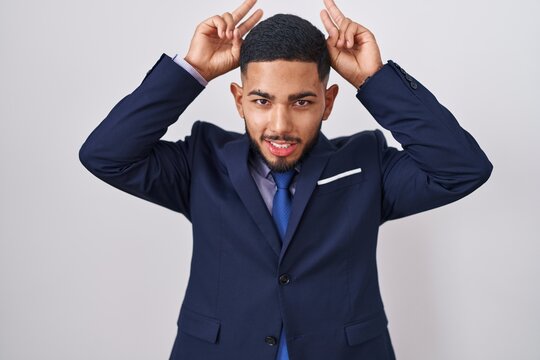 Young hispanic man wearing business suit and tie posing funny and crazy with fingers on head as bunny ears, smiling cheerful