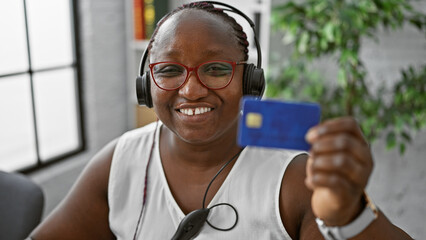 Smiling african american woman boss enjoying work at the office, managing finances with credit card, wearing headset for customer service support