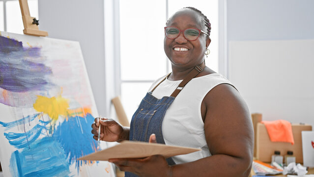 Confident, smiling african american woman artist joyfully drawing in art studio with her brush and canvas, embracing creativity.